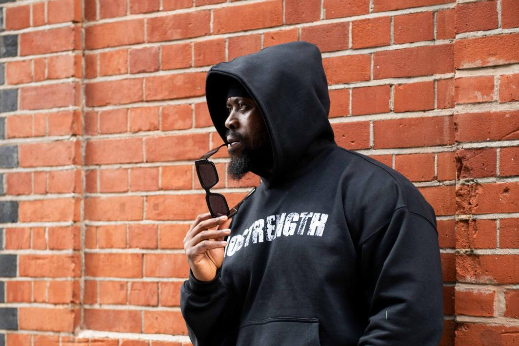 Person wearing a black hoodie with text, standing against a brick wall.